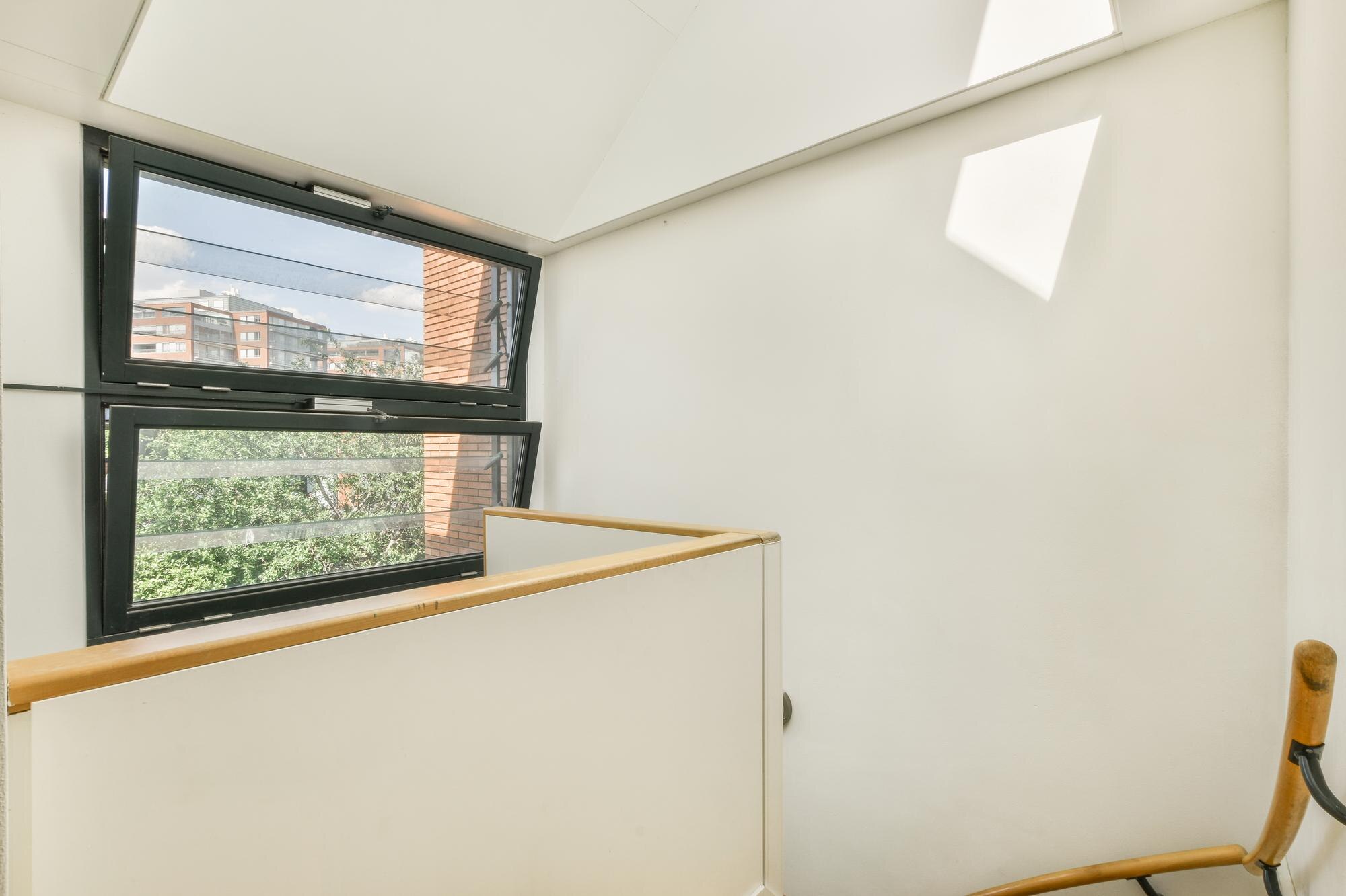 Bright stairwell/landing with white walls, wooden handrail, and a large tilted window showing a brick building outside.