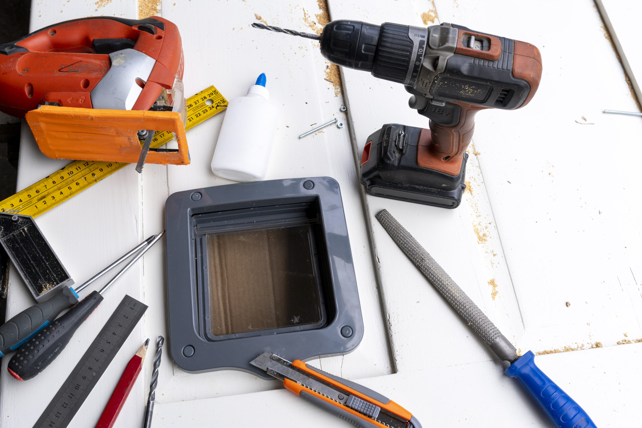 assorted tools on a white wooden work surface orange cordless drill tape measure rulers and a handheld device with a screen