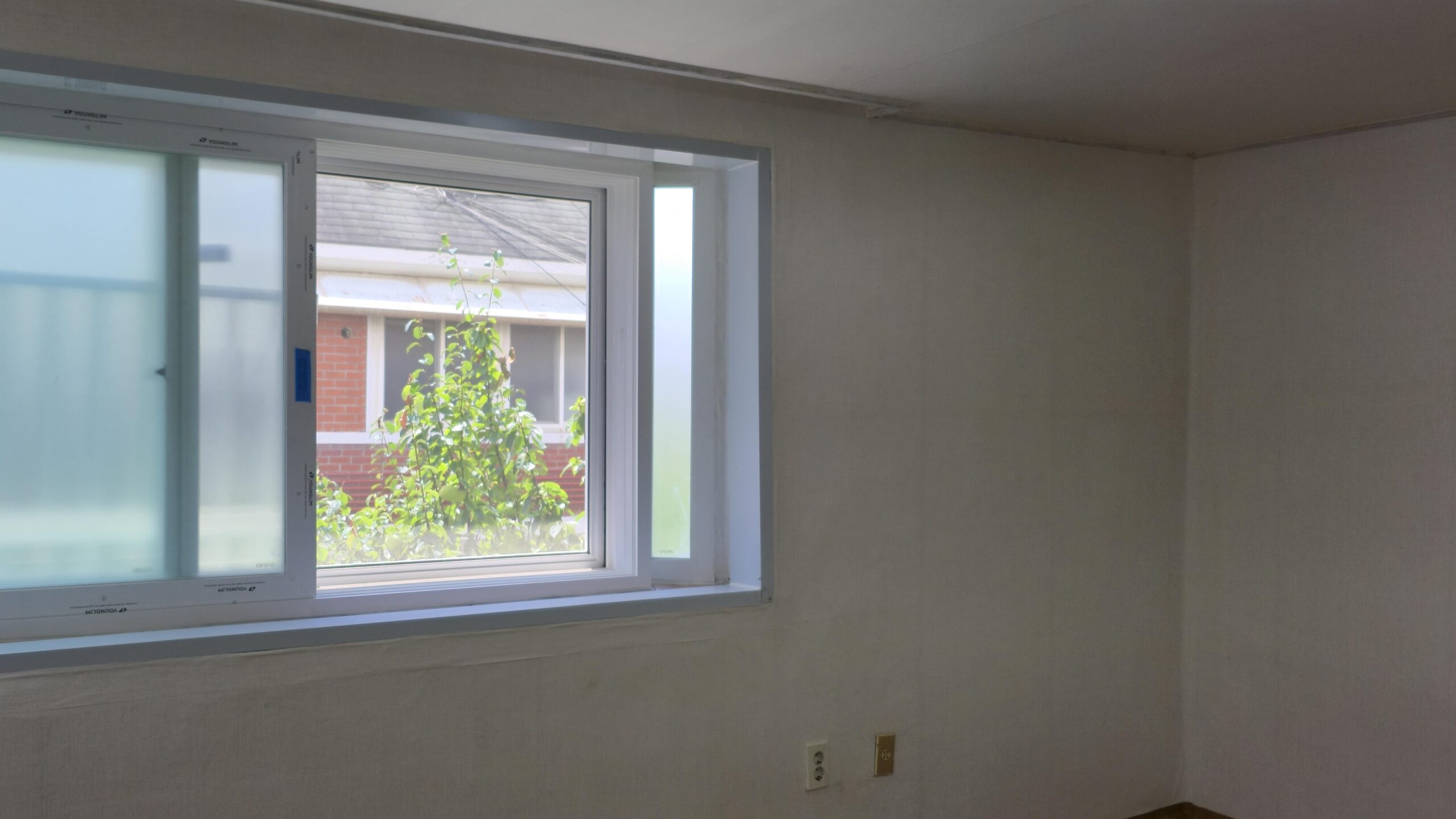 interior room with a large white framed window outside green foliage and a brick building are visible against a pale wall interior