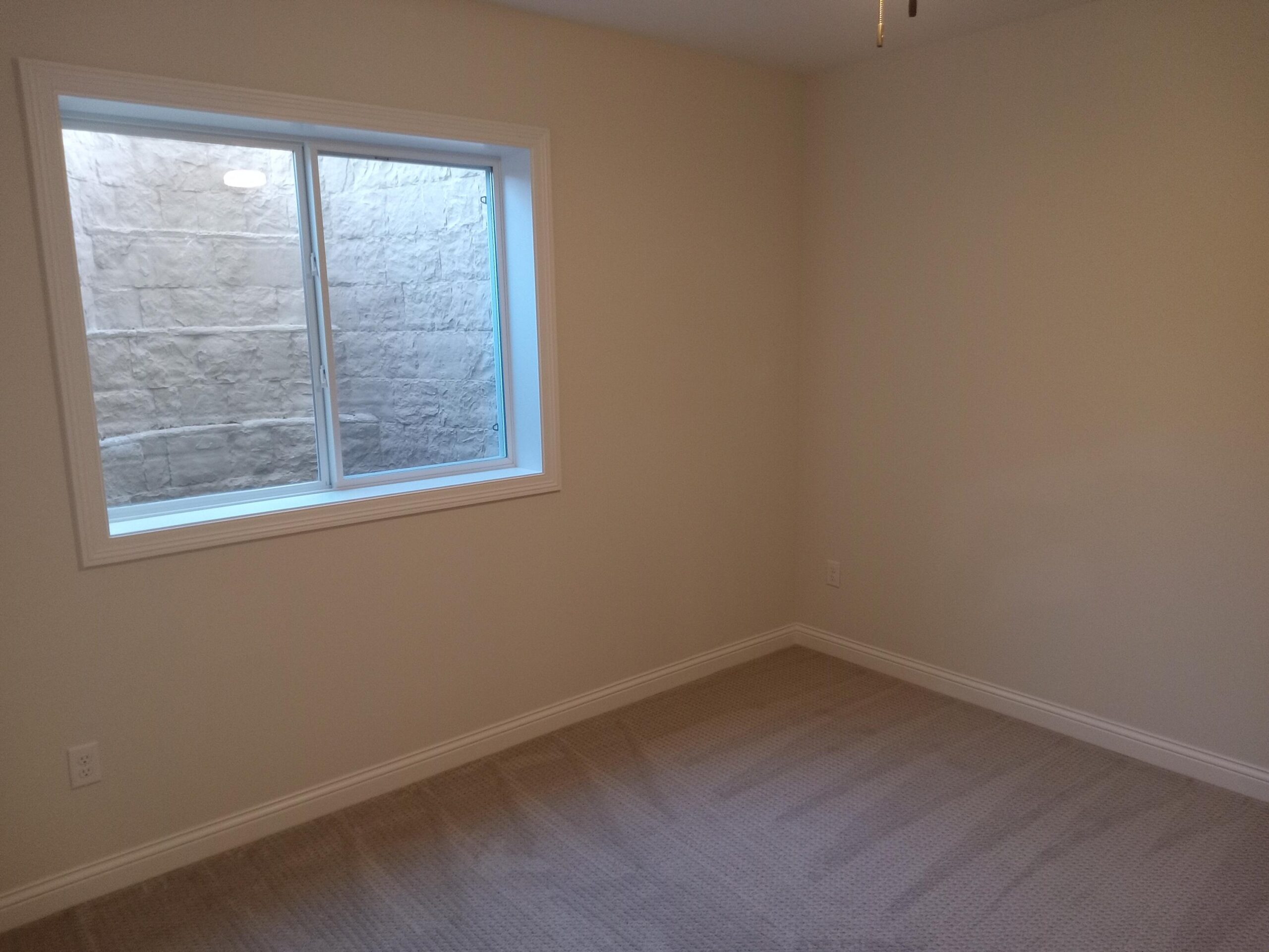 Corner of an empty beige room with a white-framed window showing a stone exterior wall outside and beige carpeted floor.