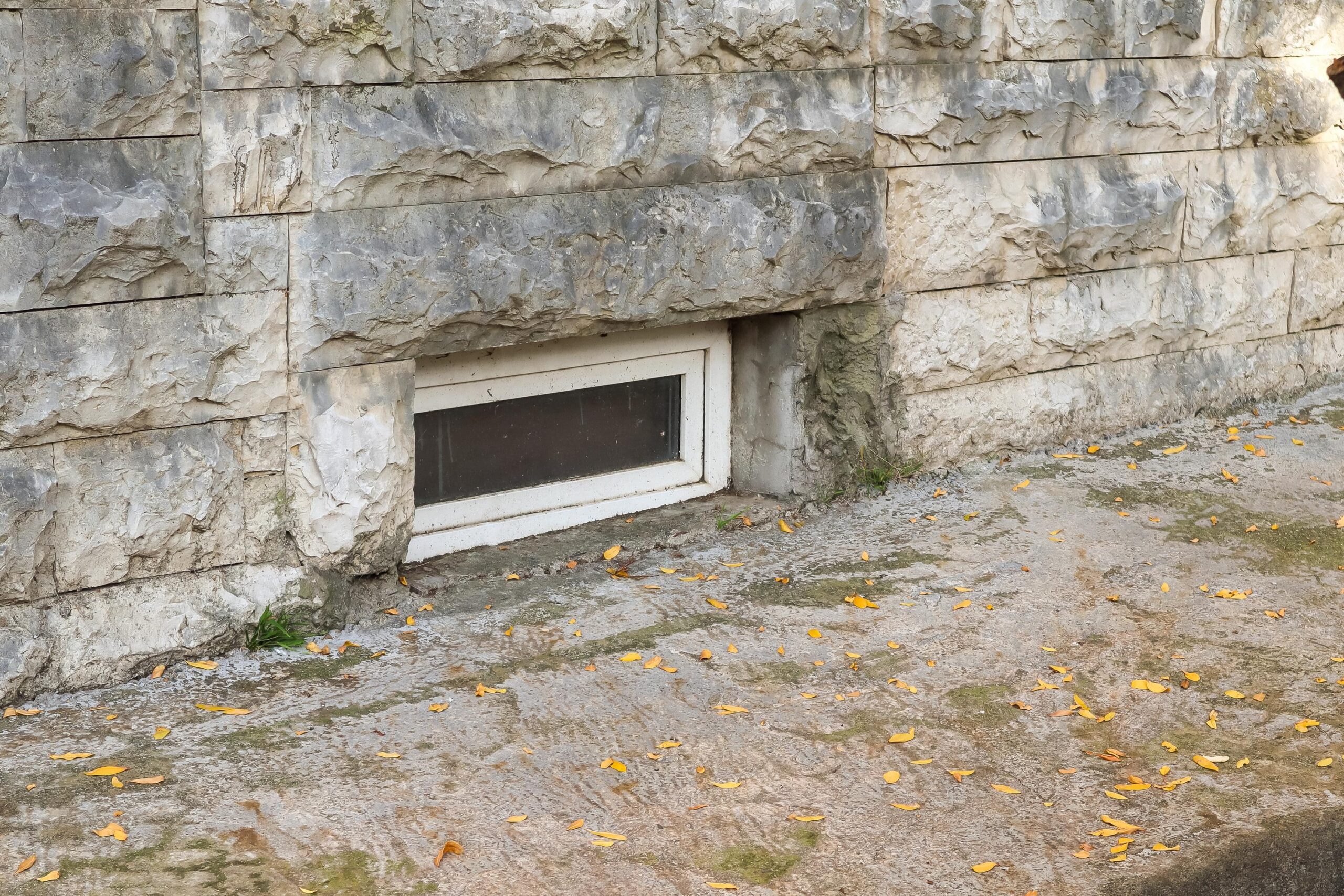 Stone stone wall with a small rectangular basement window, ground scattered with yellow leaves.