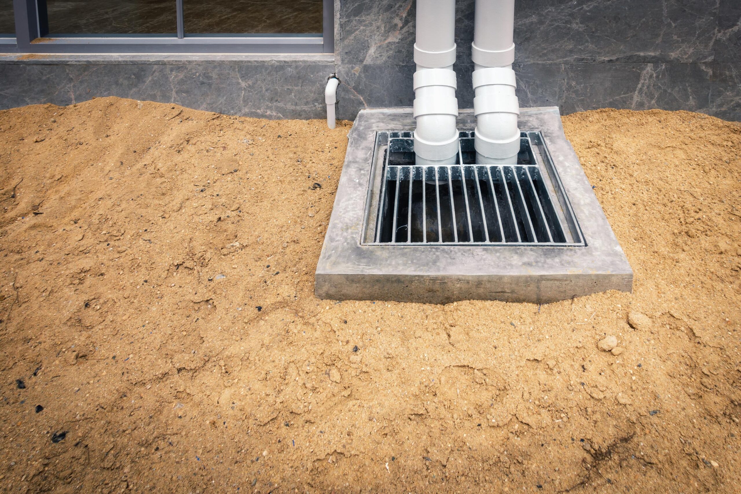 Open grated drain with two vertical PVC pipes next to a building, surrounded by a dirt mound.