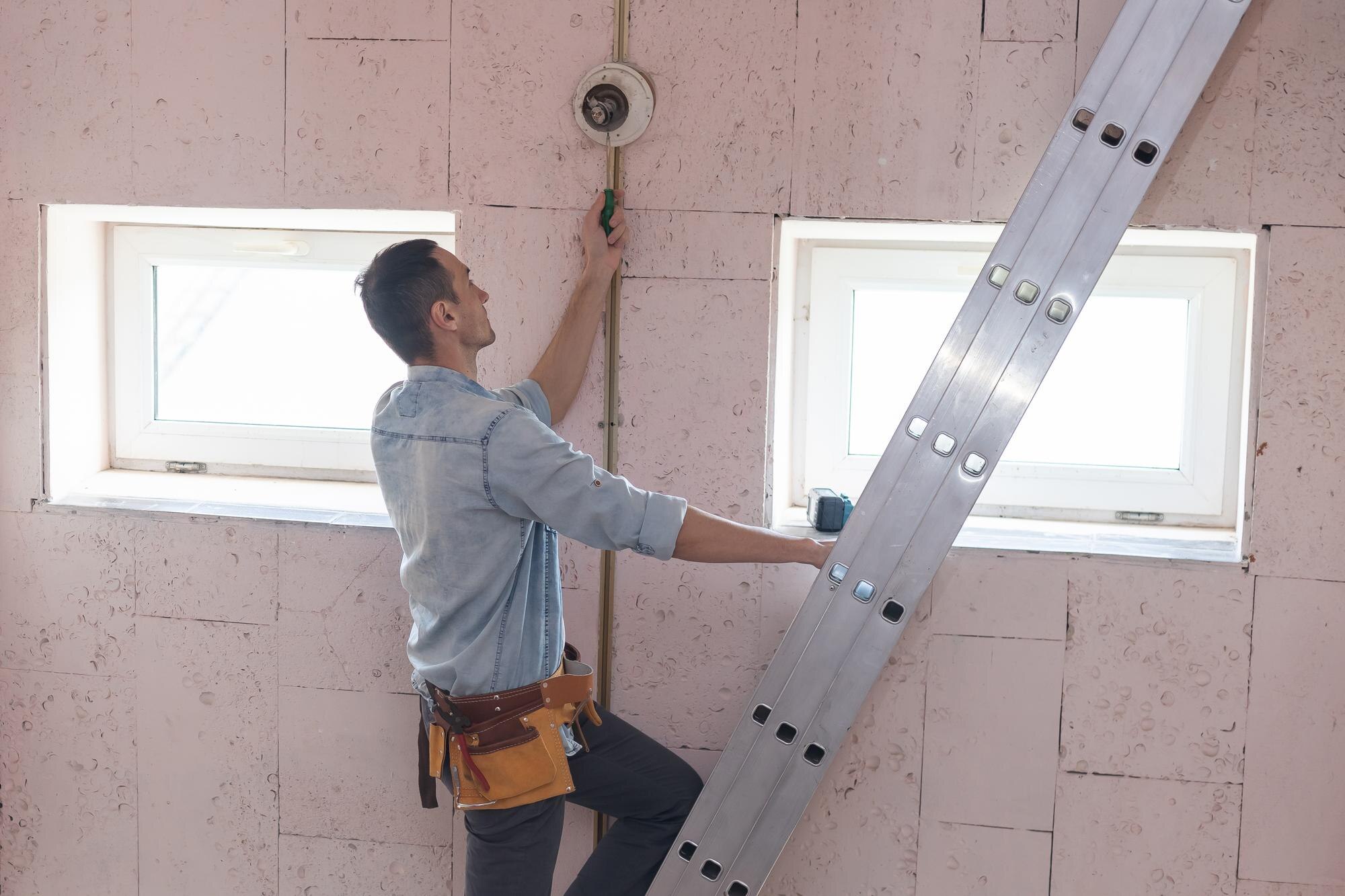 Construction worker standing on a ladder, wiring a wall in a room under renovation, with two windows nearby.