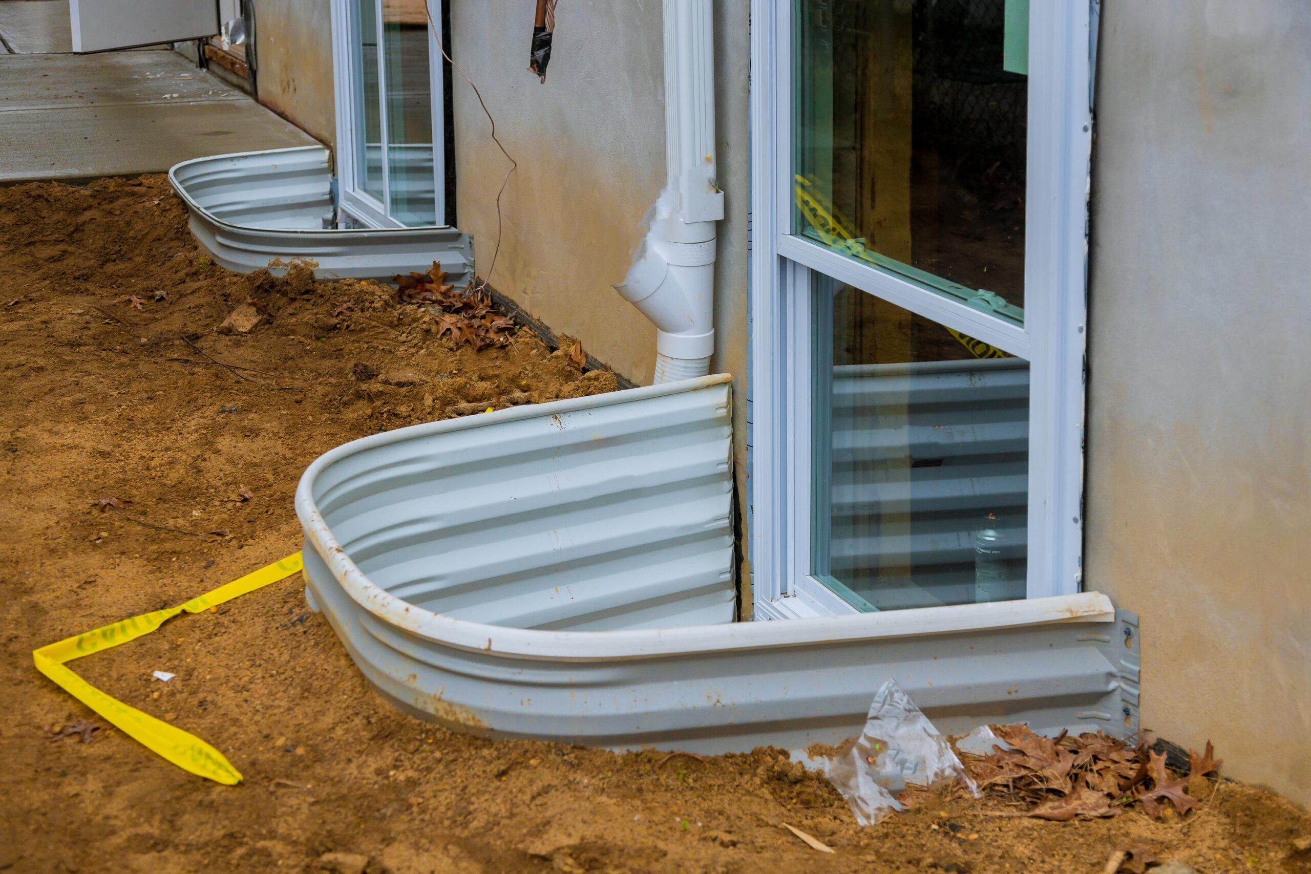 Building exterior under construction with dirt piled around a curved white window well and a downspout; yellow caution tape on the ground nearby.