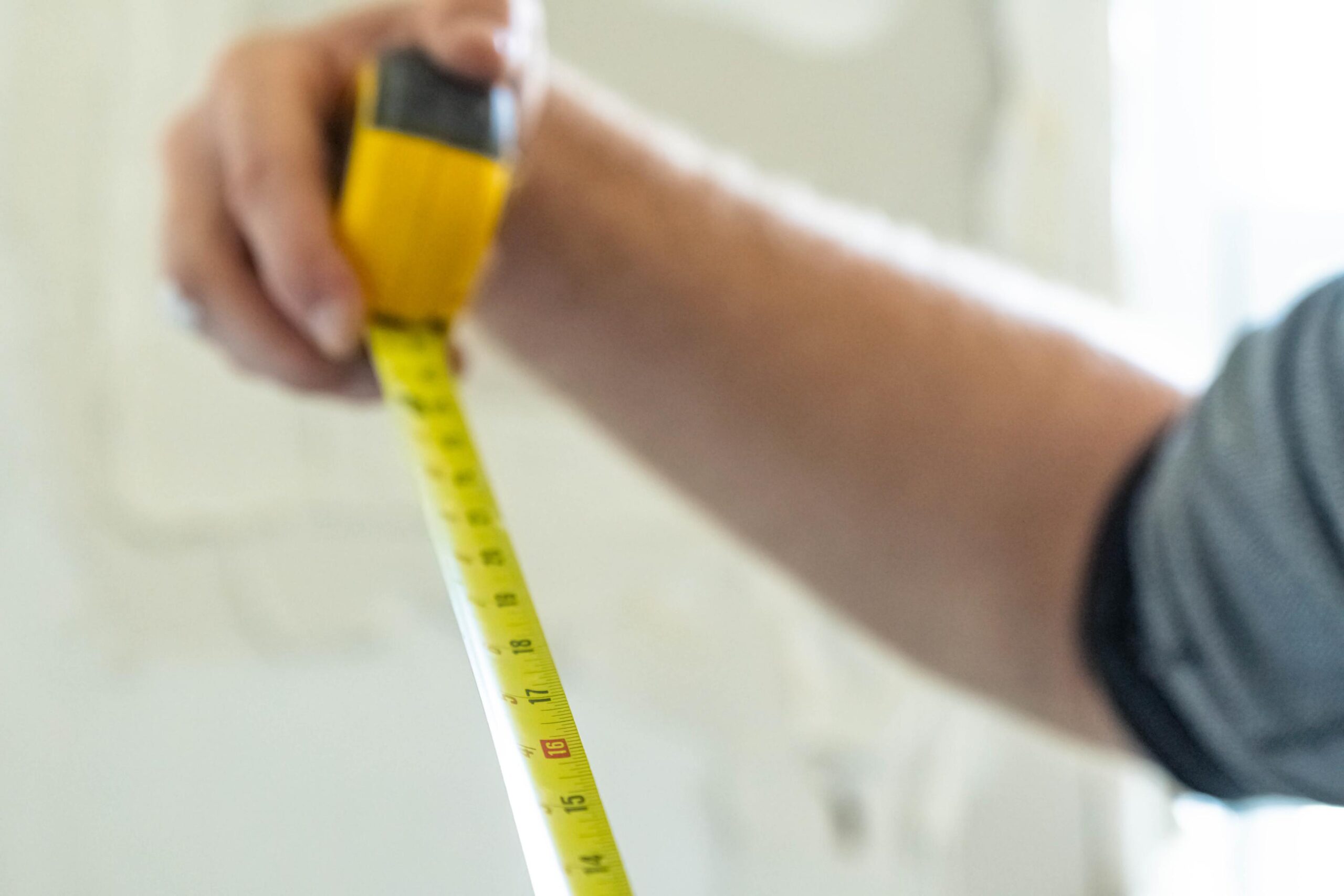 Close-up of a hand holding a yellow measuring tape extended diagonally across the frame, blurred background.