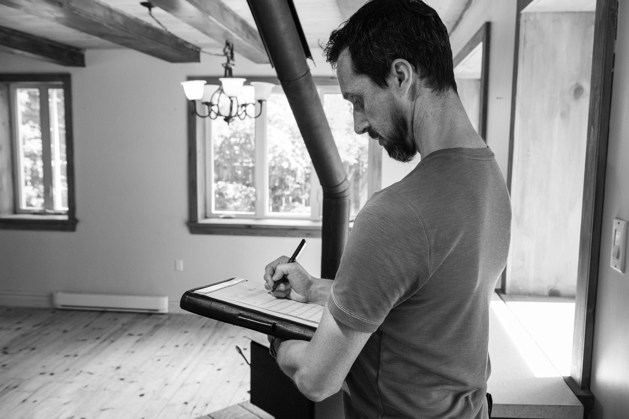 Man in a t‑shirt writes on a clipboard indoors, near large windows letting in light.