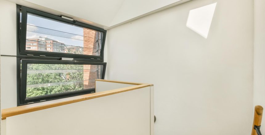 Bright stairwell/landing with white walls, wooden handrail, and a large tilted window showing a brick building outside.