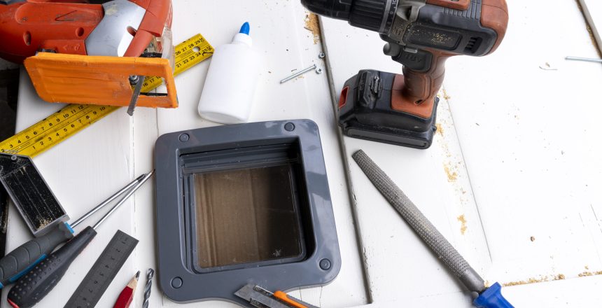 Assorted tools on a white wooden work surface: orange cordless drill, tape measure, rulers, and a handheld device with a screen.