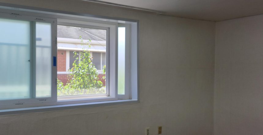 Interior room with a large white-framed window; outside, green foliage and a brick building are visible against a pale wall interior.