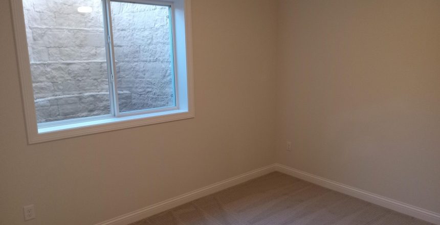Corner of an empty beige room with a white-framed window showing a stone exterior wall outside and beige carpeted floor.