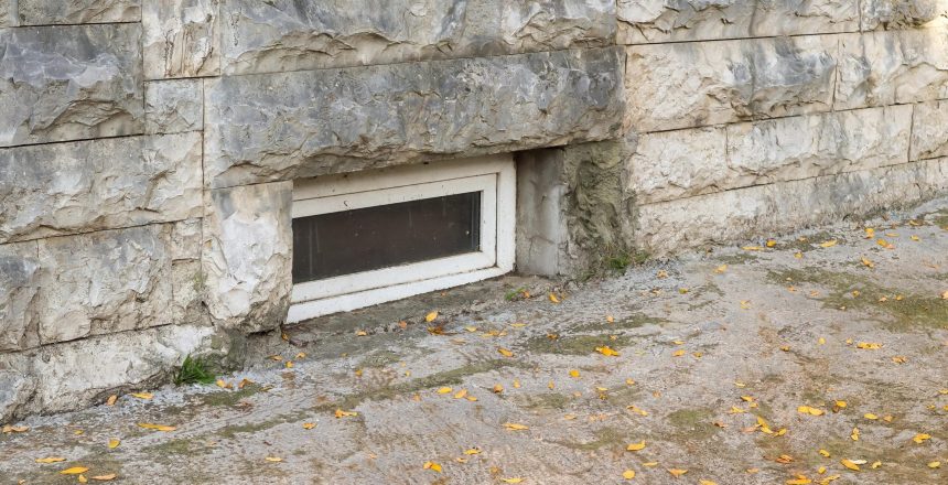 Stone stone wall with a small rectangular basement window, ground scattered with yellow leaves.