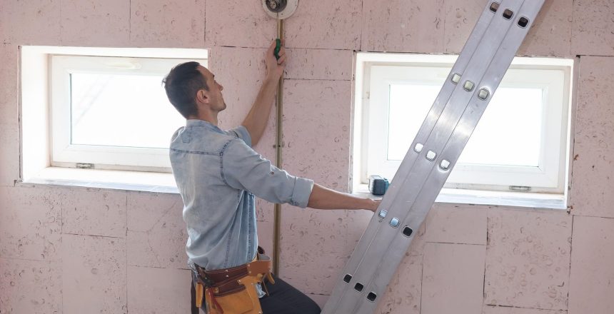 Construction worker standing on a ladder, wiring a wall in a room under renovation, with two windows nearby.