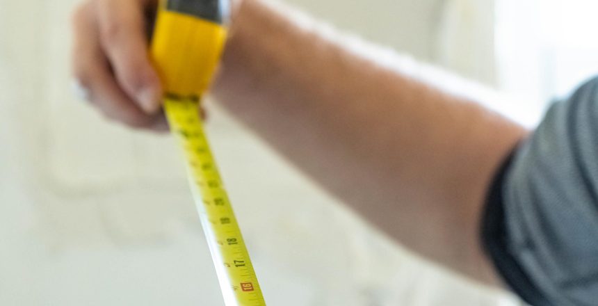 Close-up of a hand holding a yellow measuring tape extended diagonally across the frame, blurred background.
