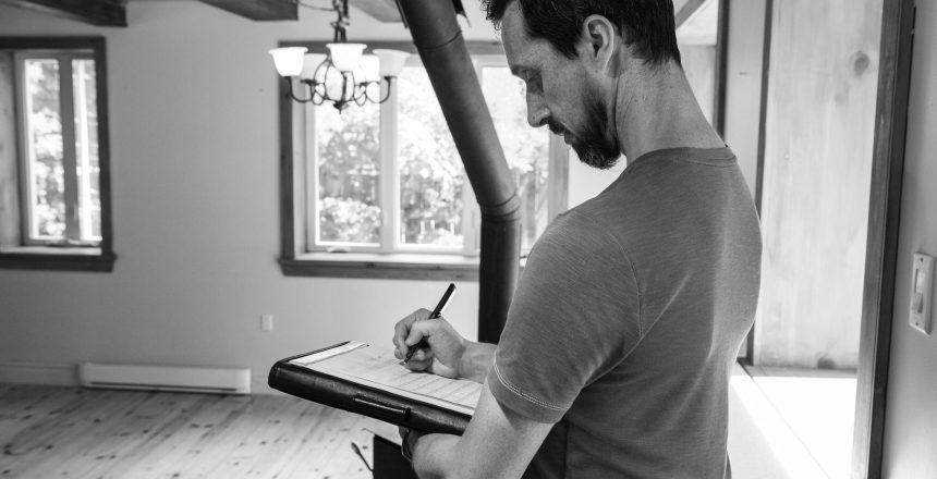 Man in a t‑shirt writes on a clipboard indoors, near large windows letting in light.