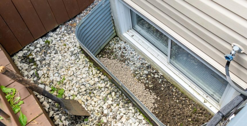 Gravel and soil mixture fills a window well, bordered by decorative stones and a shovel, illustrating effective landscaping and drainage techniques
