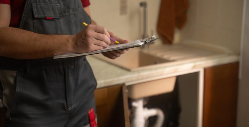 Experienced maintenance professional conducting thorough kitchen sink plumbing inspection, carefully documenting findings with clipboard and detailed notes for comprehensive service evaluation